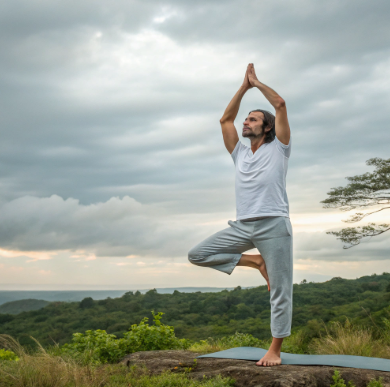 Man doing yoga in tree pose on a mat in nature, surrounded by greenery and cloudy skies.