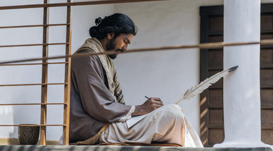 Man in traditional attire sitting on a veranda, writing with a quill pen on paper, focused and thoughtful.