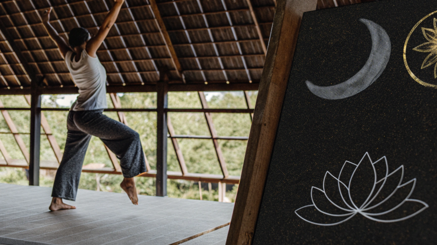 Woman practicing yoga indoors under a thatched roof, with a chalkboard showing a crescent moon and lotus flower design in the foreground.